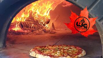 Un horno de pizza con una pizza en su interior, fuego encendido y un logo sobre una hoja.