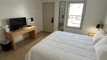 A bedroom with a bed, a desk with a TV, a lamp, a trash bin, and a window.