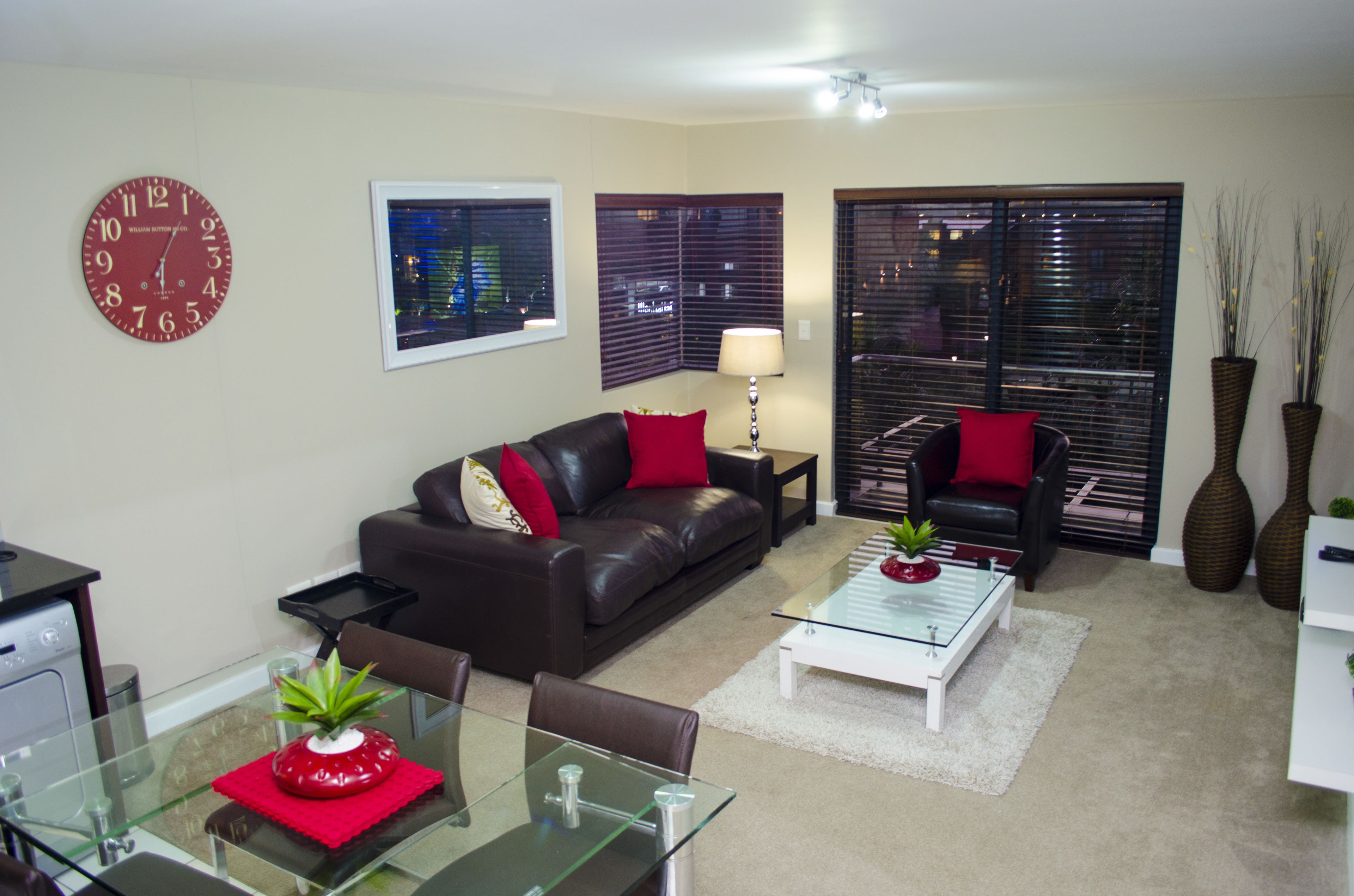 A living room with a brown leather sofa, a glass coffee table, and a red clock on the wall.
