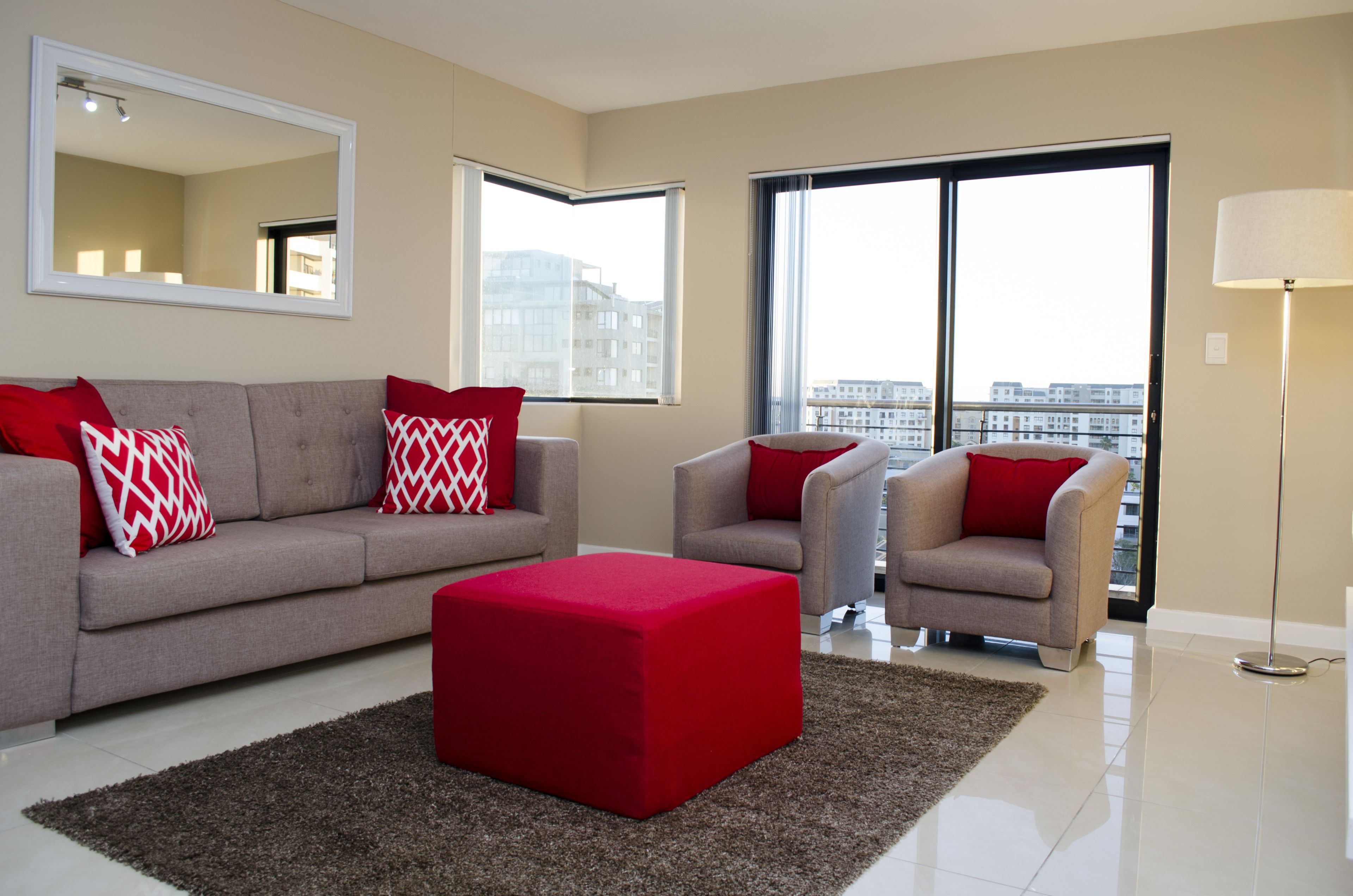A modern living room with a grey sofa, two armchairs, a red ottoman, and a large mirror.