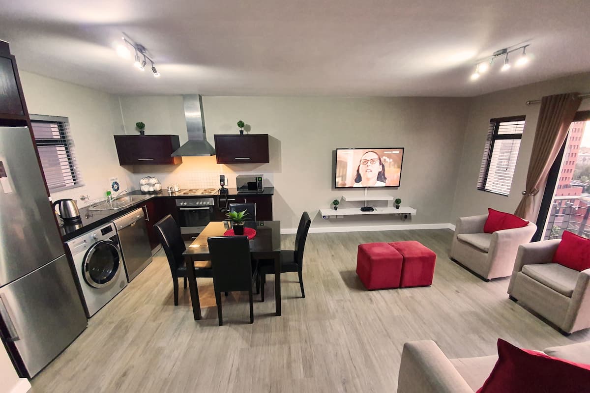 A modern apartment with a kitchen, dining area, and living room. It includes a washing machine, a dining table with chairs, a TV, and a sofa with red cushions.