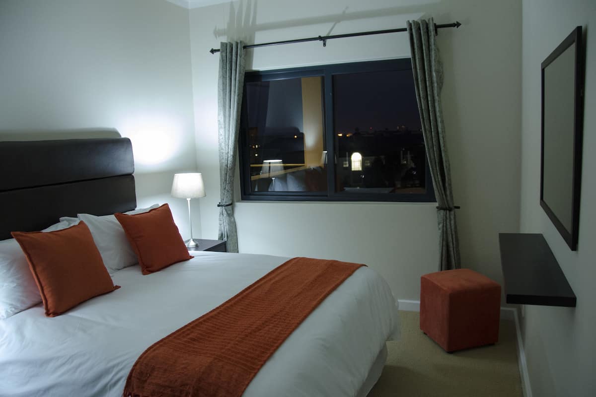 A hotel room with a large bed, a nightstand with a lamp, a window with curtains, a small ottoman, and a framed picture on the wall.