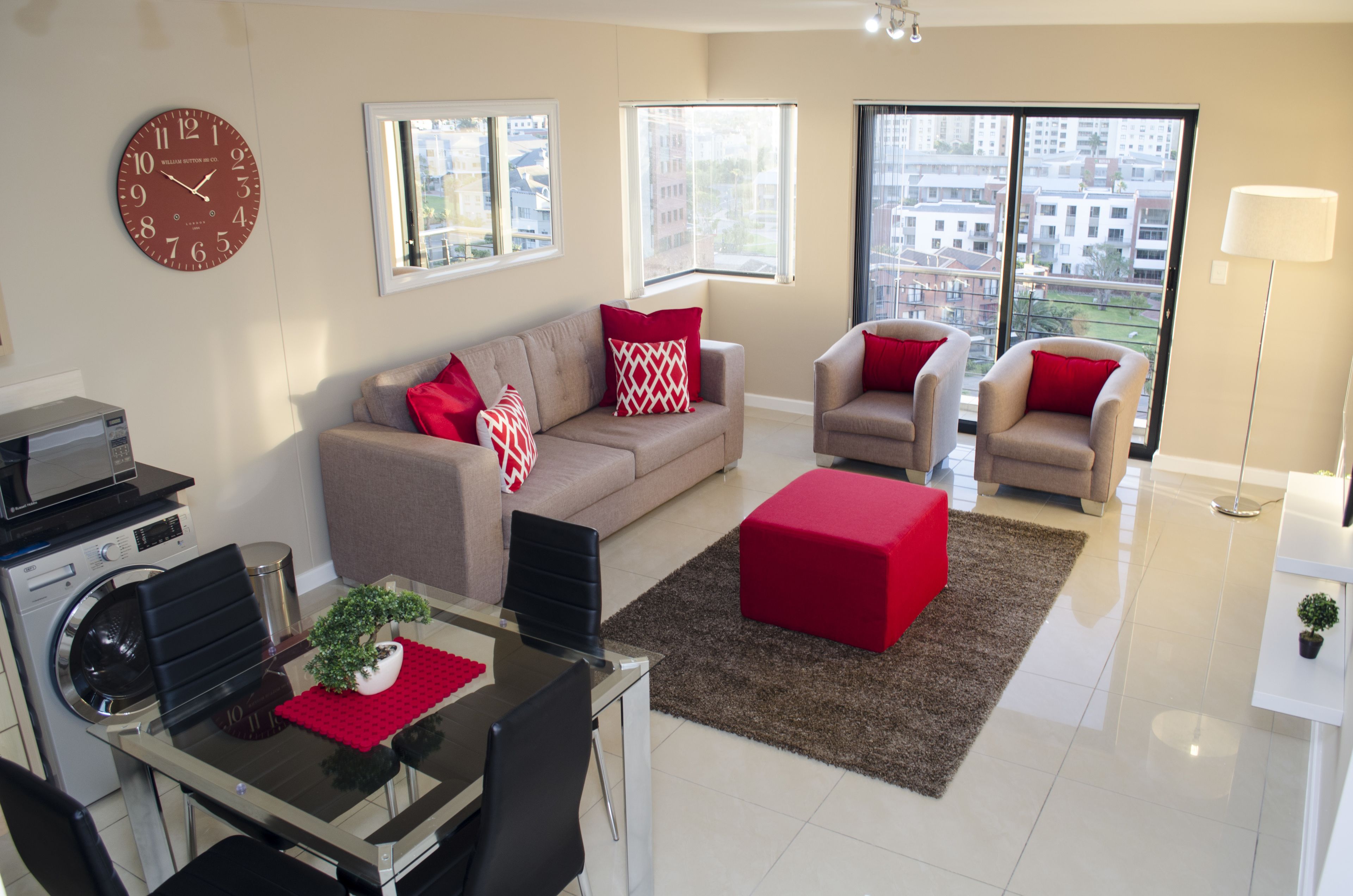 A modern living room with a sofa, armchairs, a dining table, and a red ottoman. There is a large window providing a view of the city.