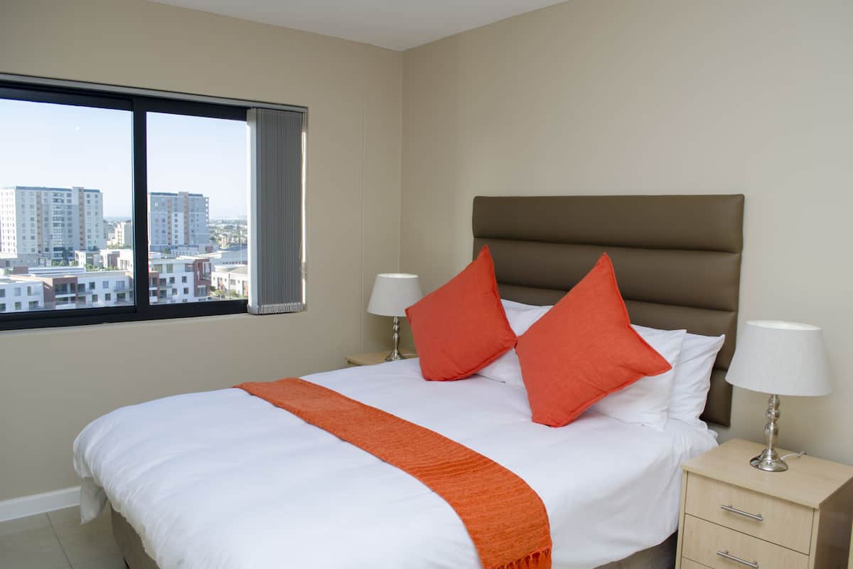 A hotel room with a large bed, two pillows, a nightstand, a lamp, and a window with a city view.