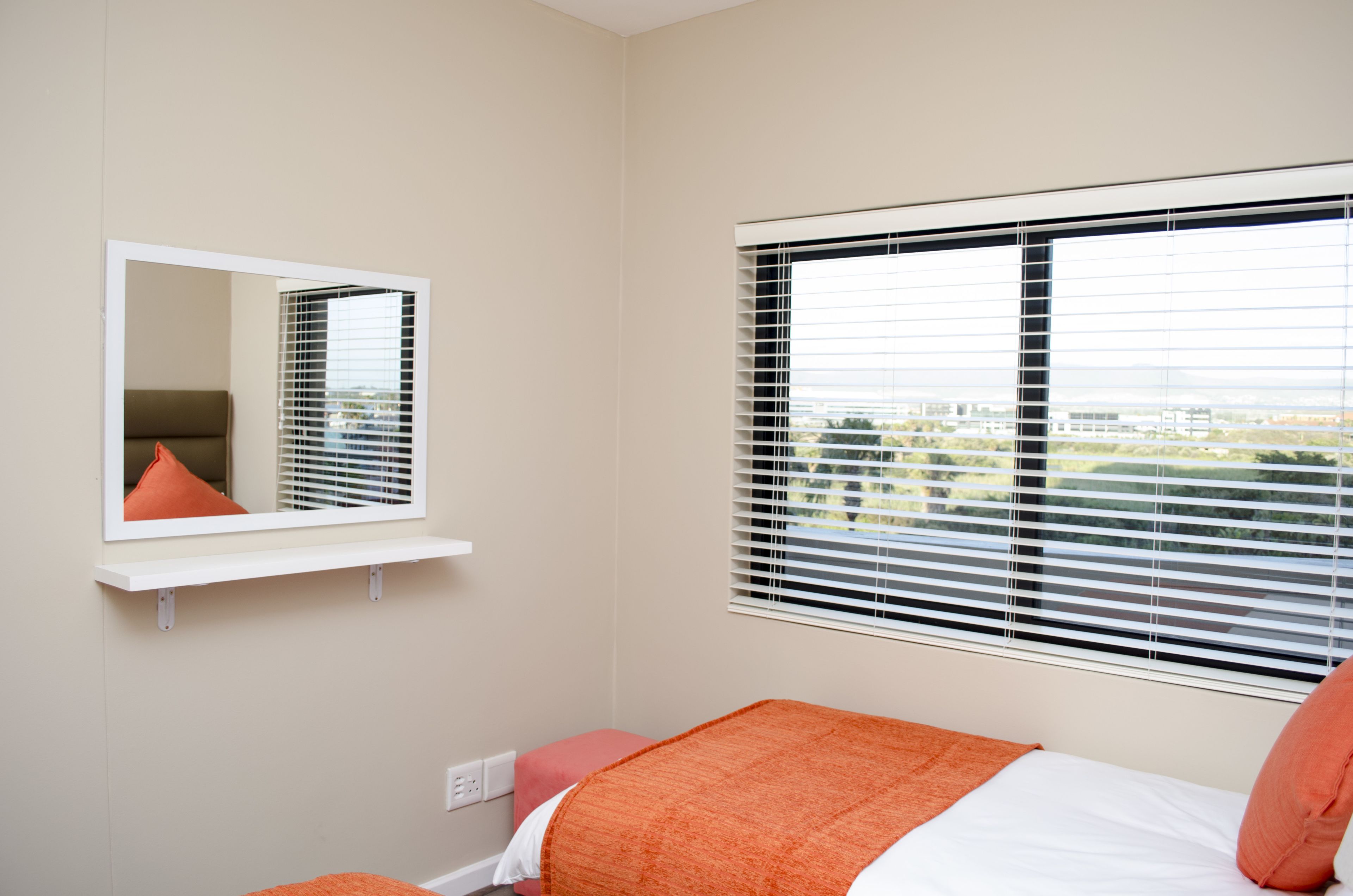 A bedroom with a bed, a window with blinds, a mirror, and a view of the city.