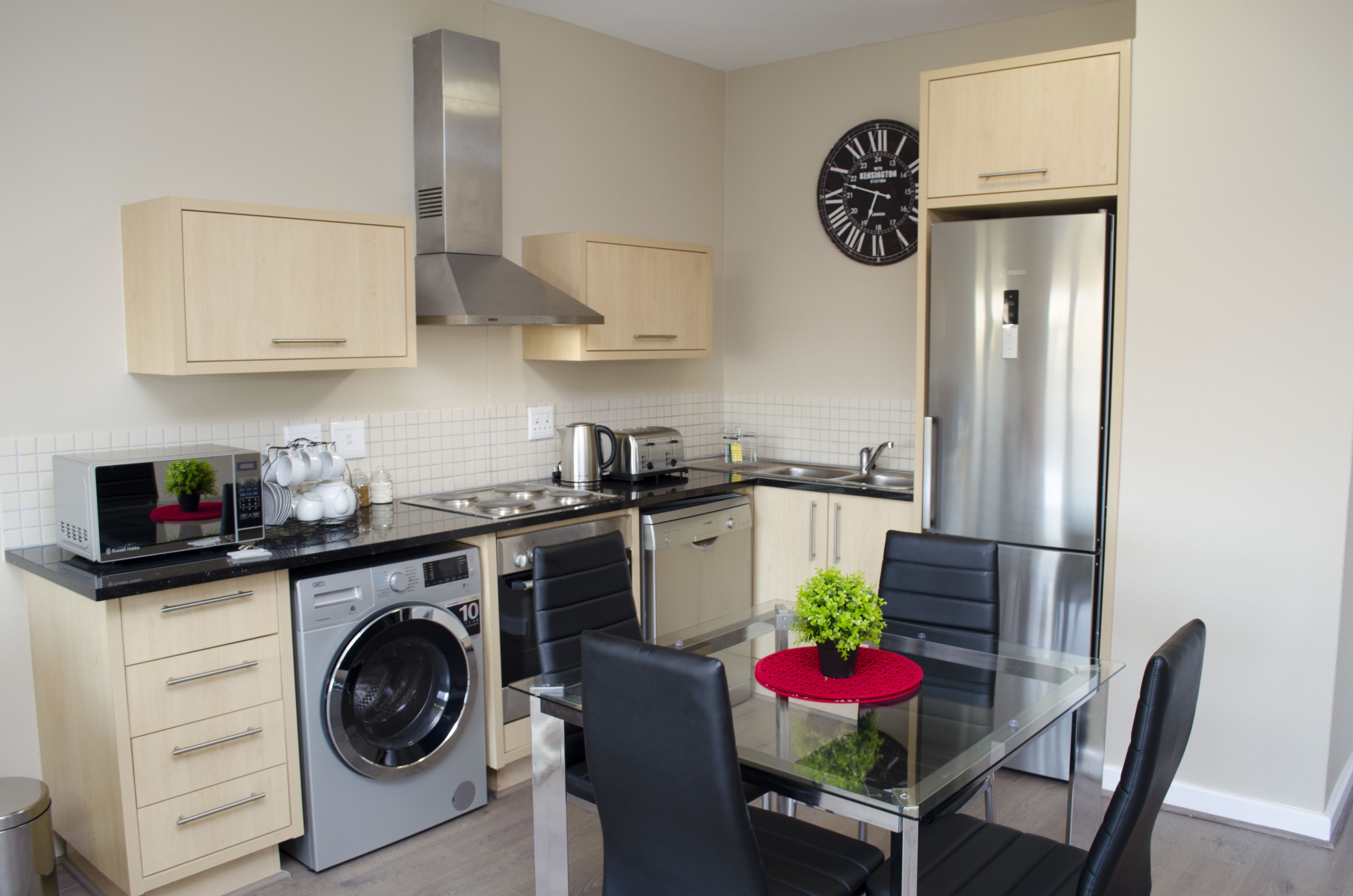 A modern kitchen with a dishwasher, washing machine, stainless steel appliances, and a dining area with a glass table and chairs.