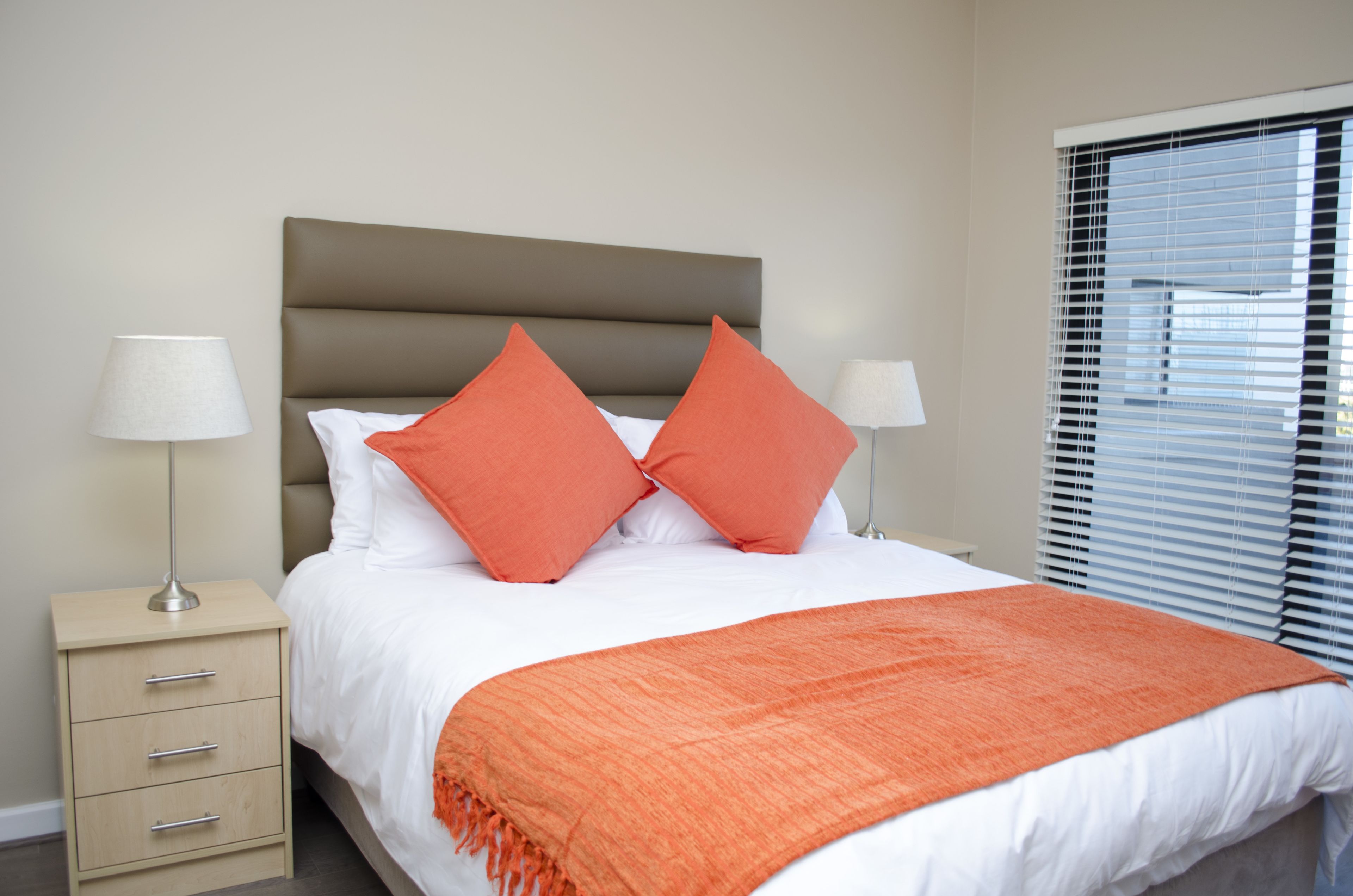 Una cama bien hecha con ropa de cama blanca y naranja, una mesita de noche color beige con una lámpara y una ventana con persianas.