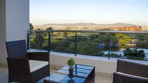 Een balkon met rieten meubels, een glazen tafel en uitzicht op een rivier met boten en gebouwen in de verte.