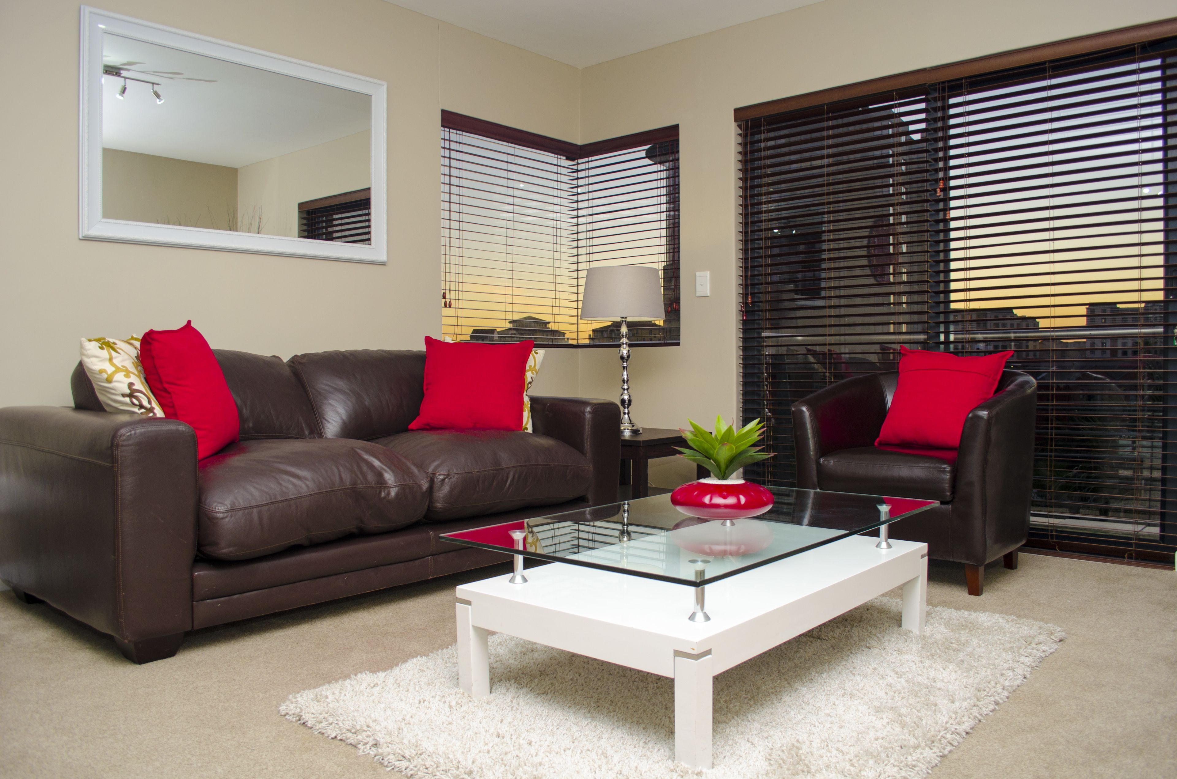 A living room with a brown leather sofa, a glass coffee table, and two red pillows. There is a mirror on the wall and a window with blinds.