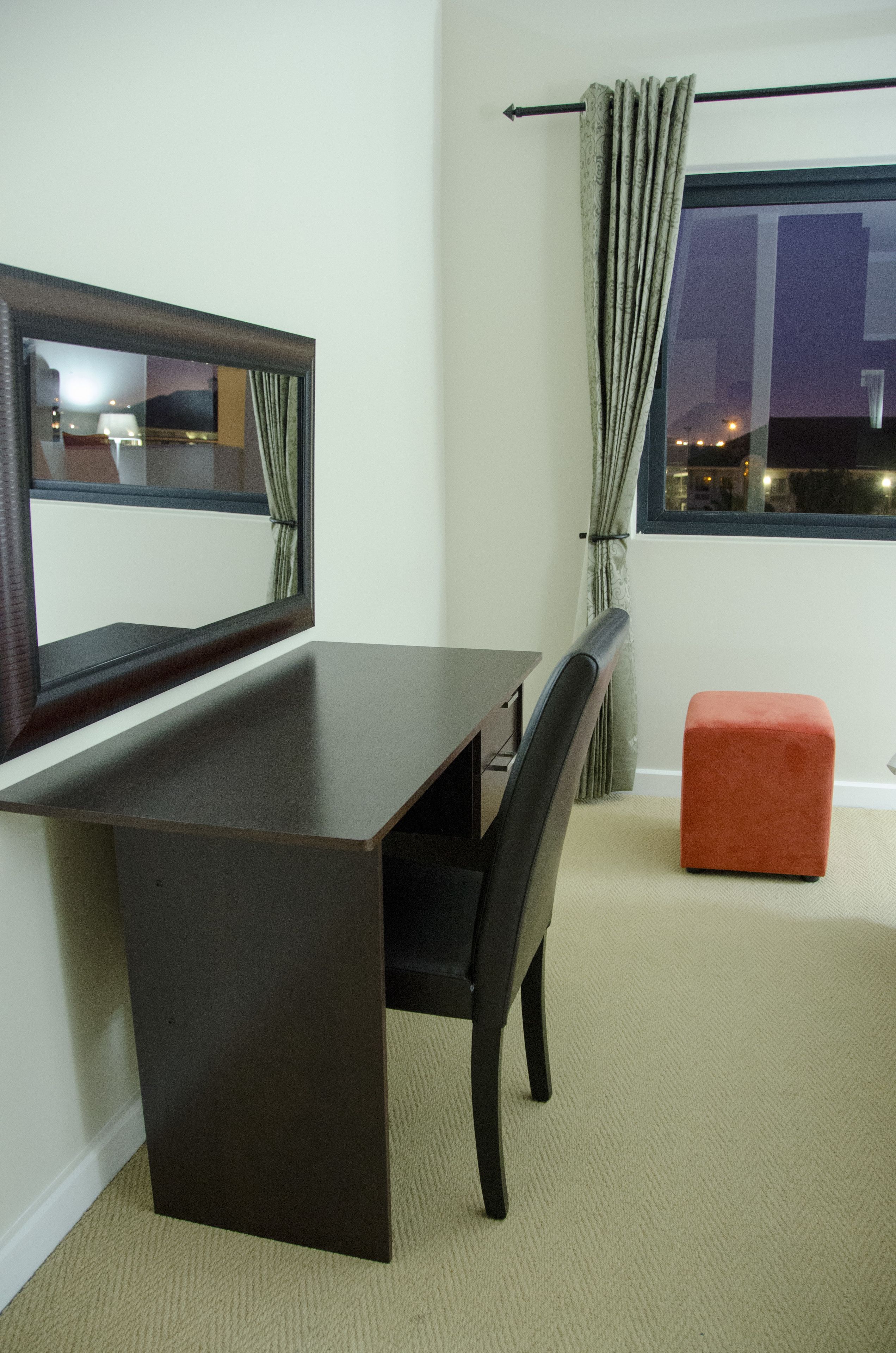A room with a dark wooden desk, a chair, a small orange ottoman, a mirror, and a window with curtains.