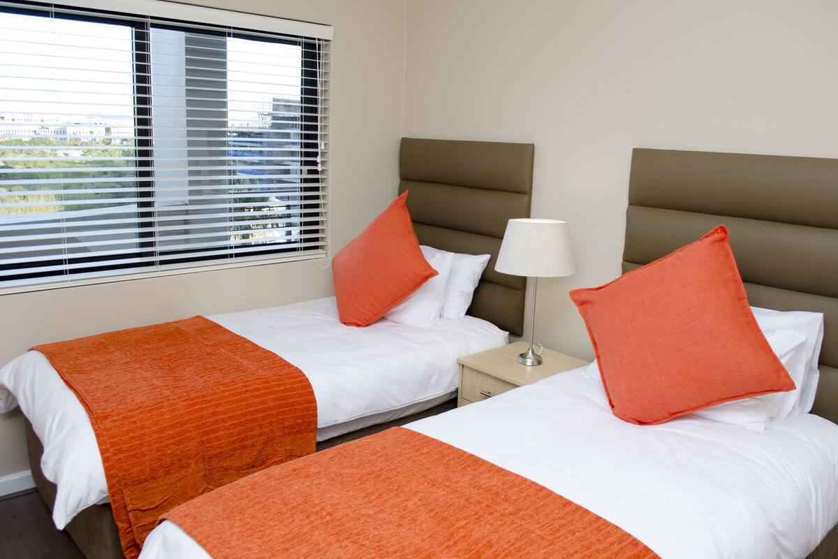 A hotel room with two beds, each with white bedding and orange throw blankets. There is a nightstand with a lamp between the beds.