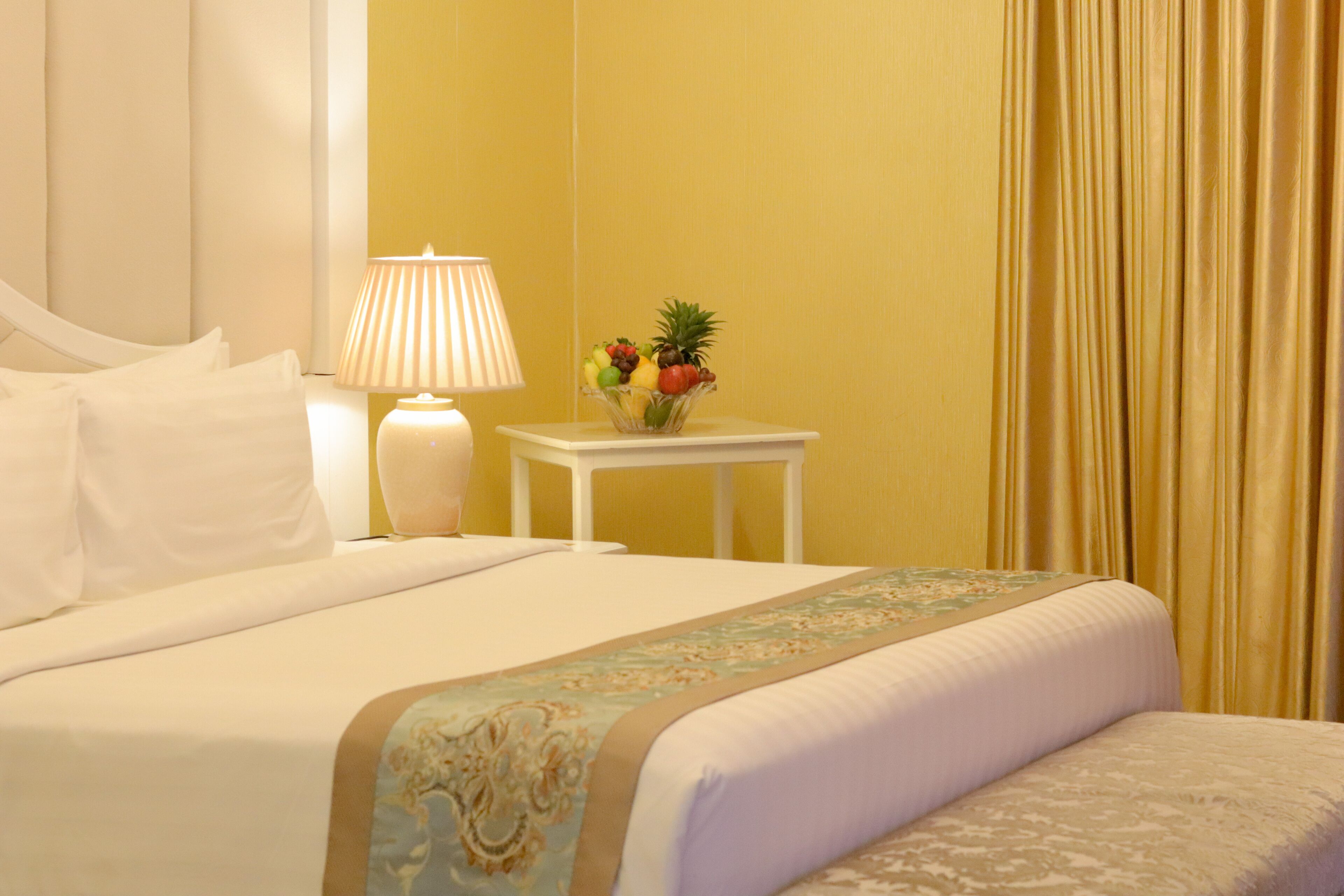 A neatly made bed with white linens, a bedside table with a lamp, and a fruit bowl.