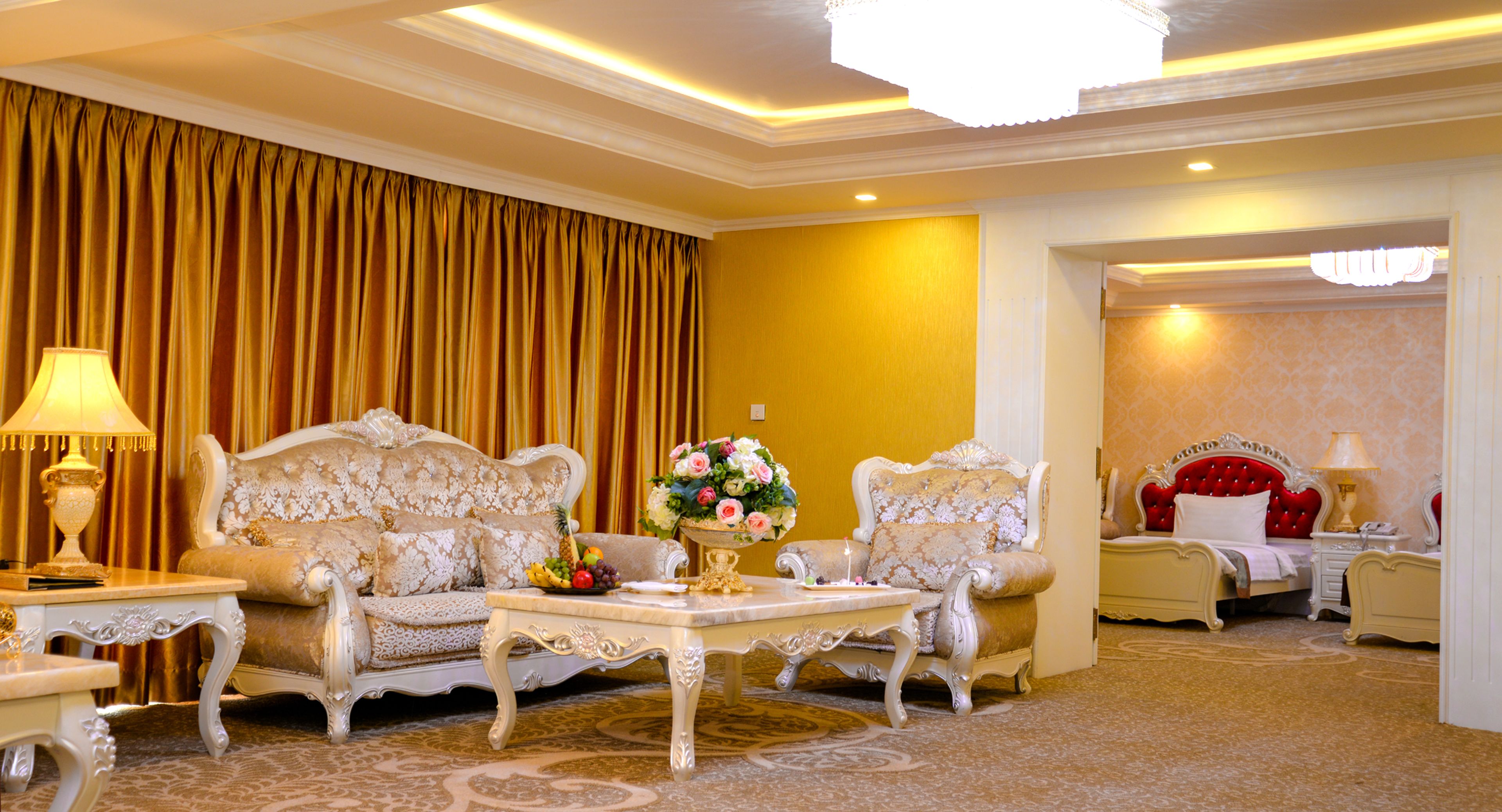 A luxurious hotel room with a classic design, featuring a large sofa, a matching armchair, a coffee table, and a bed with ornate headboard.