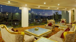 Un hall d’hôtel luxueux, doté de fauteuils confortables, d’une grande baie vitrée donnant sur la ville et d’un sol recouvert d’un tapis rouge.