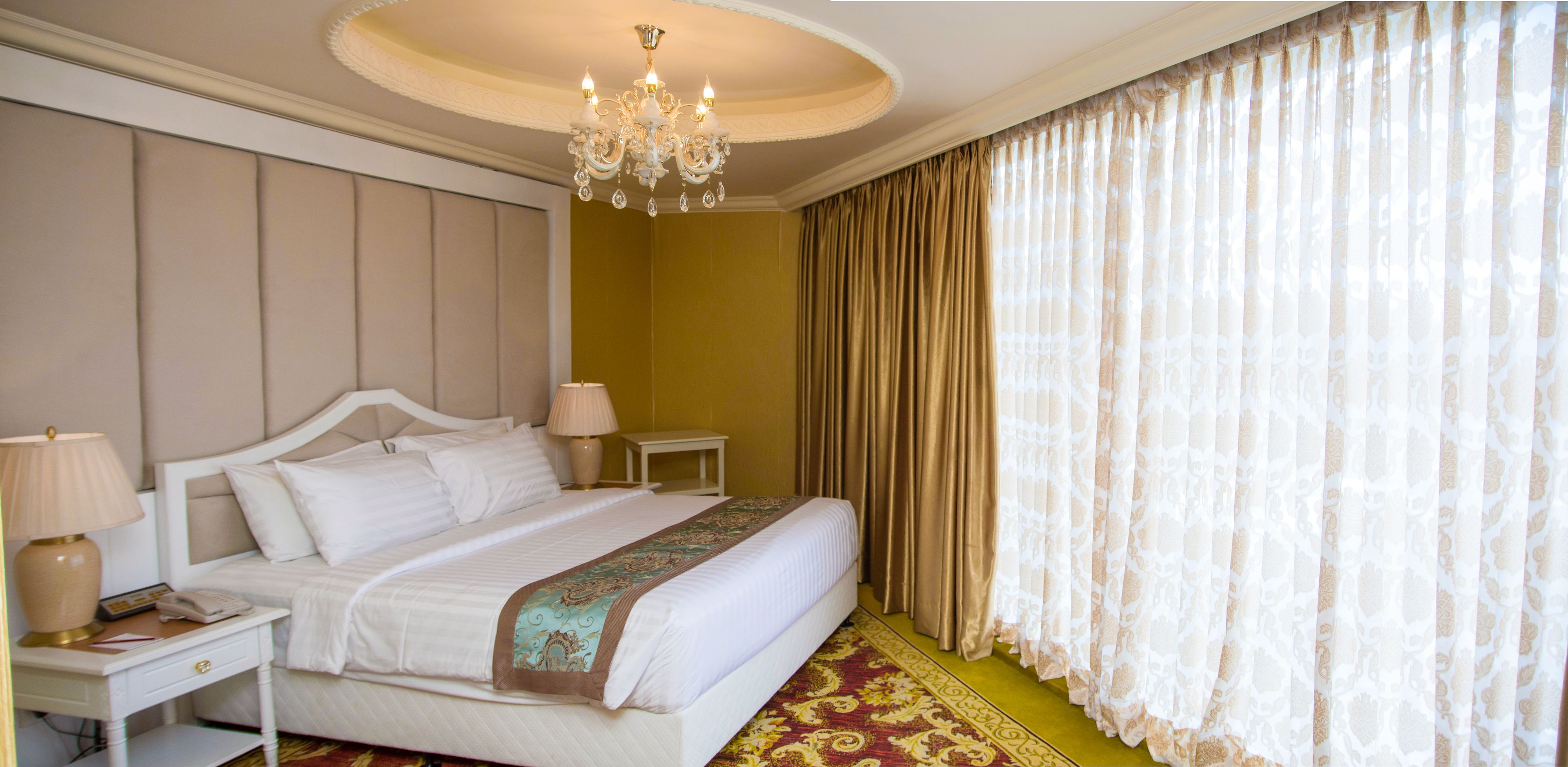 A hotel room with a large bed, a chandelier, a nightstand with a lamp, and a window with curtains.