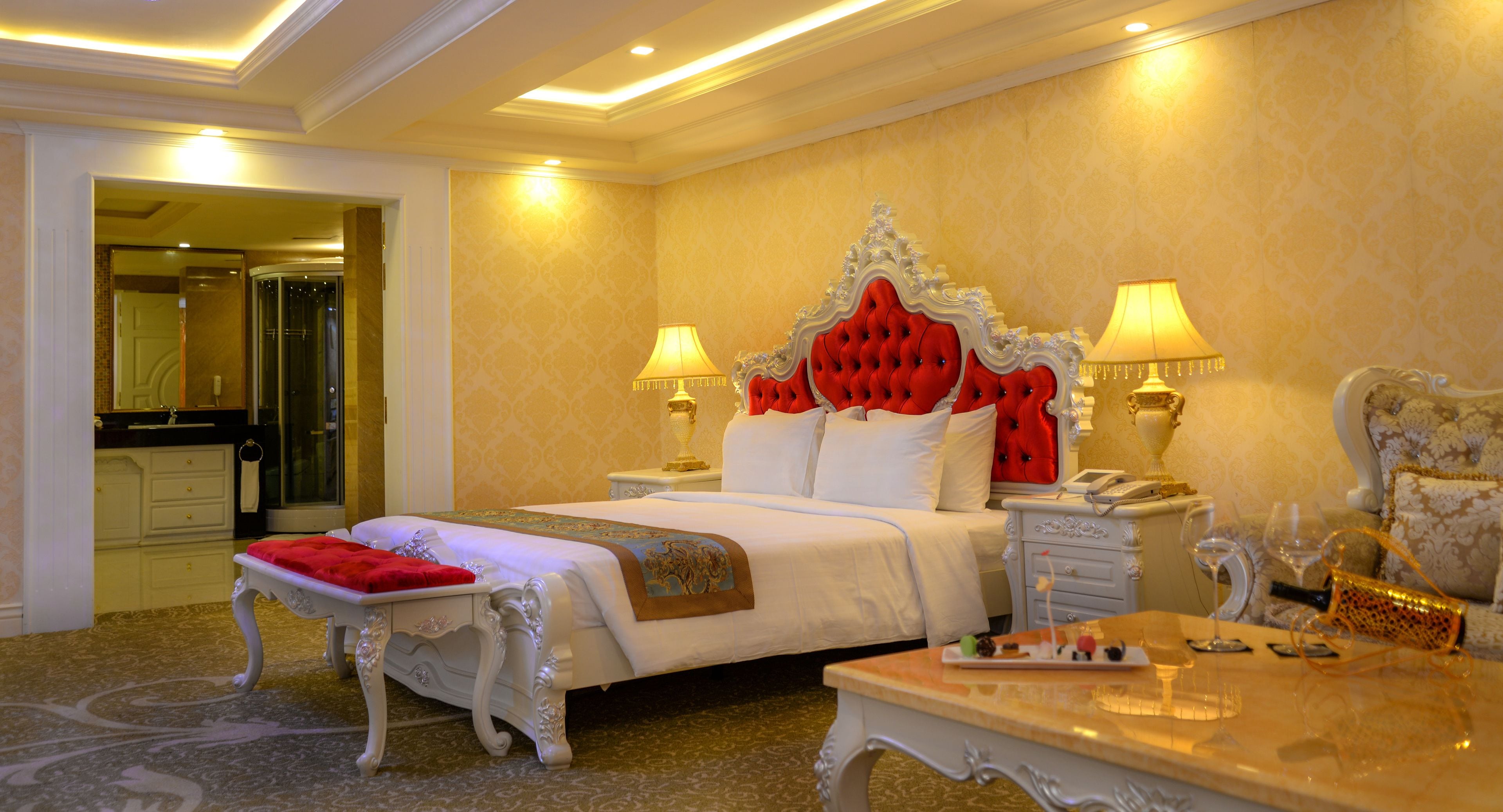 Una lujosa habitación de hotel con una cama grande, dos mesitas de noche, una mesita auxiliar, una mesa de centro de mármol y un banco tapizado en rojo.