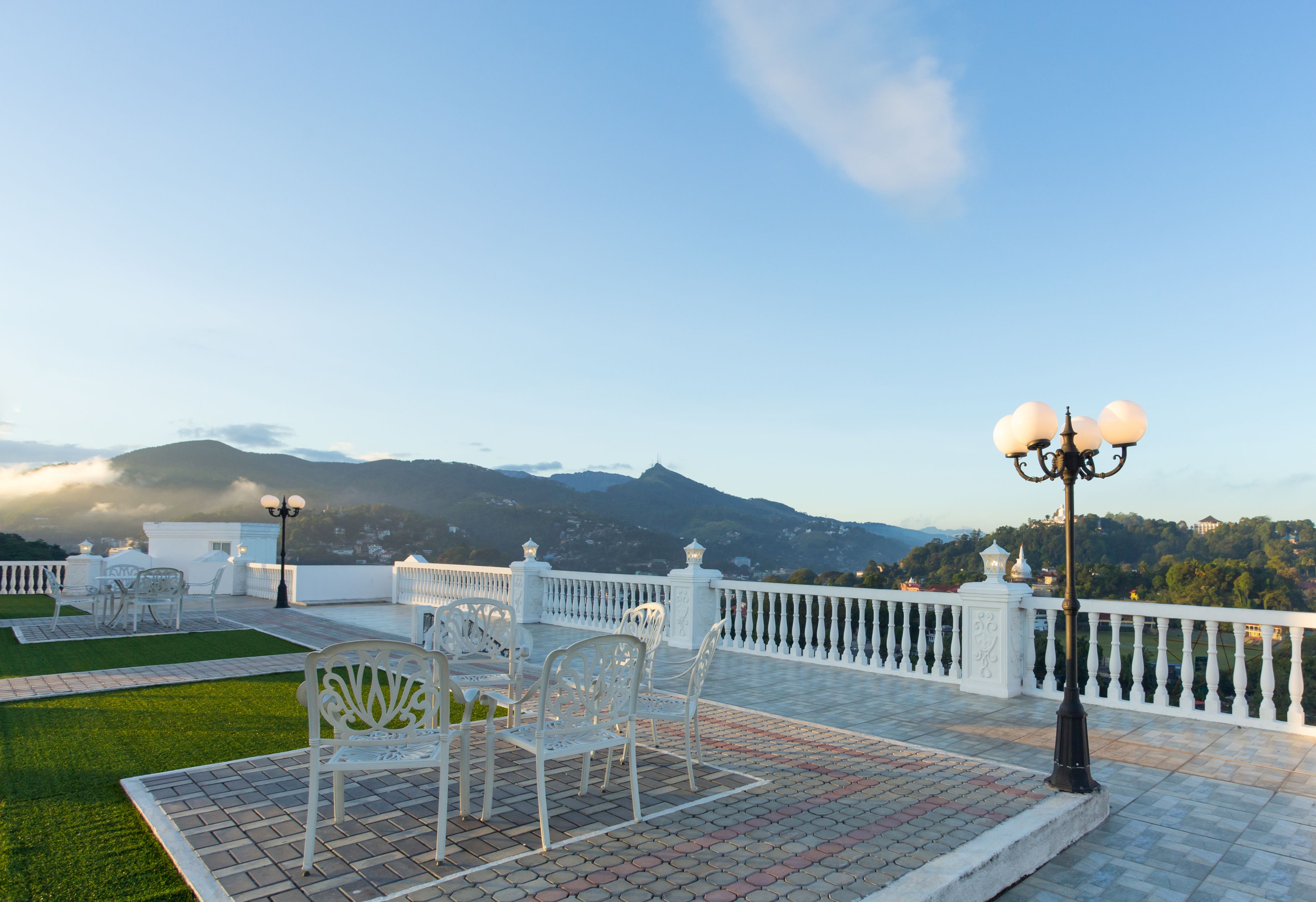 A terrace with white chairs and tables, a white railing, and a view of mountains in the background.