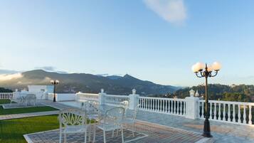 A terrace with white chairs and tables, a white railing, and a view of mountains in the background.