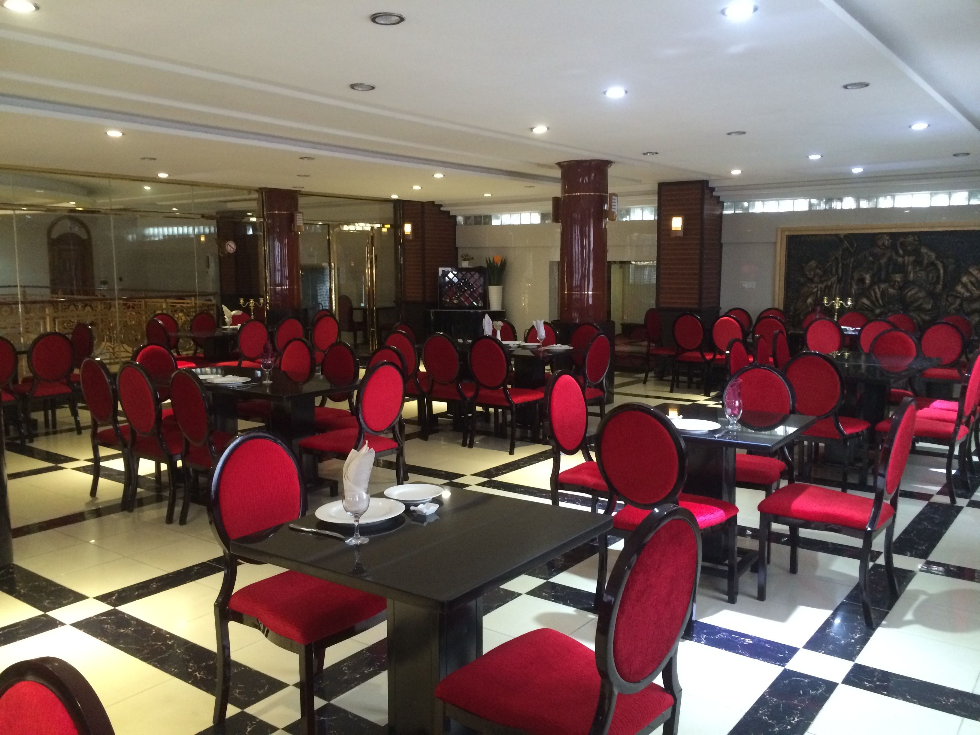 Un restaurante con sillas tapizadas de rojo y mesas negras, dispuesto en un espacio amplio con un patrón de piso a cuadros.