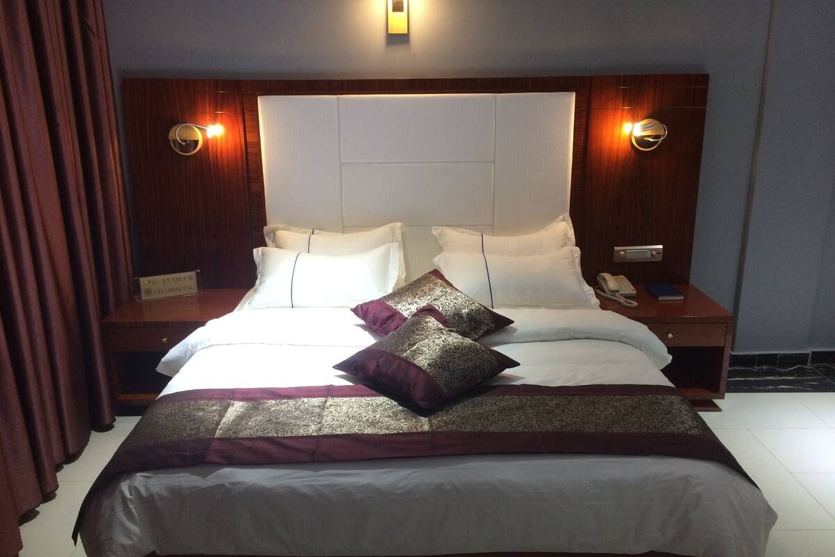 A hotel room with a large bed, two bedside tables, a nightstand, and a wall-mounted light fixture.