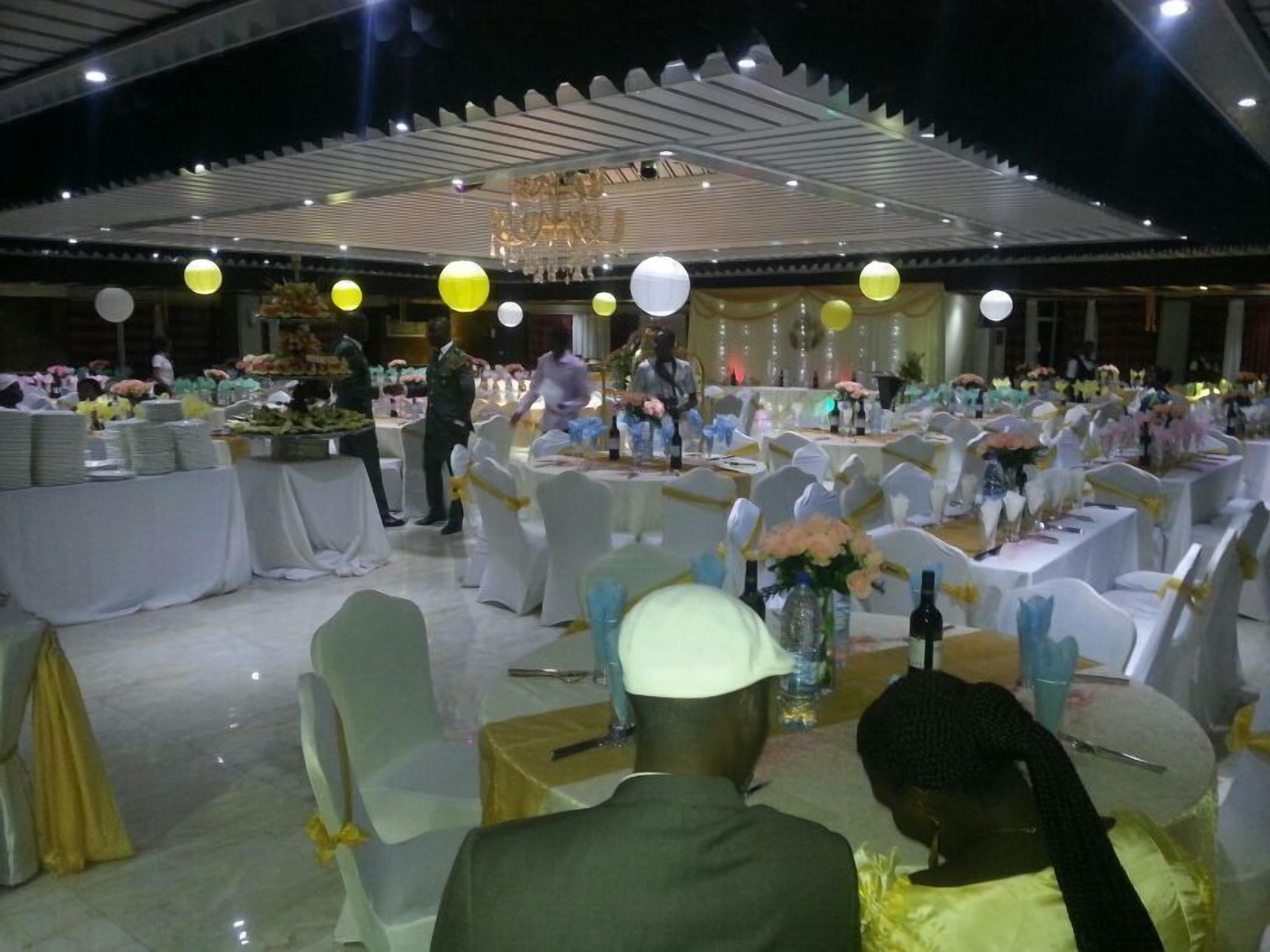 Salão de banquetes com mesas redondas preparadas para um evento formal, com toalhas brancas, cadeiras com fitas amarelas e arranjos florais.