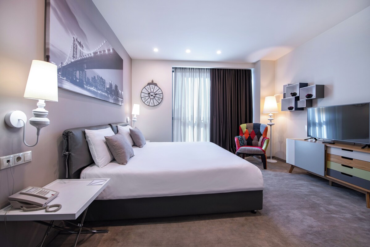 Quarto de hotel moderno com uma cama grande, uma mesa com telefone, televisão e uma cadeira colorida.