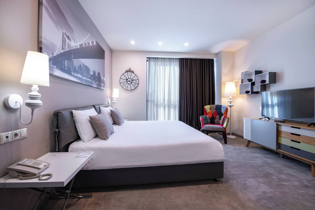 A modern hotel room with a large bed, a desk with a telephone, a television, and a colorful chair.