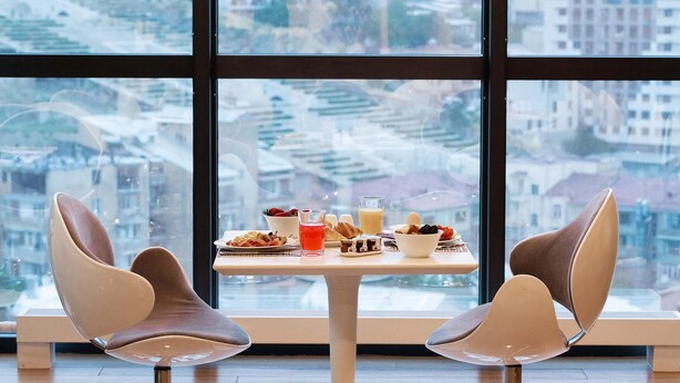 Ein Essbereich mit Tisch für zwei Personen, ausgestattet mit verschiedenen Frühstücksangeboten und einem Glas Orangensaft. Moderne Stühle und ein großes Fenster mit Blick auf eine Stadtlandschaft.