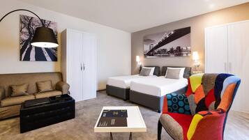 A hotel room with a sofa, two beds, a colorful armchair, a table with a tray, and artwork on the walls.