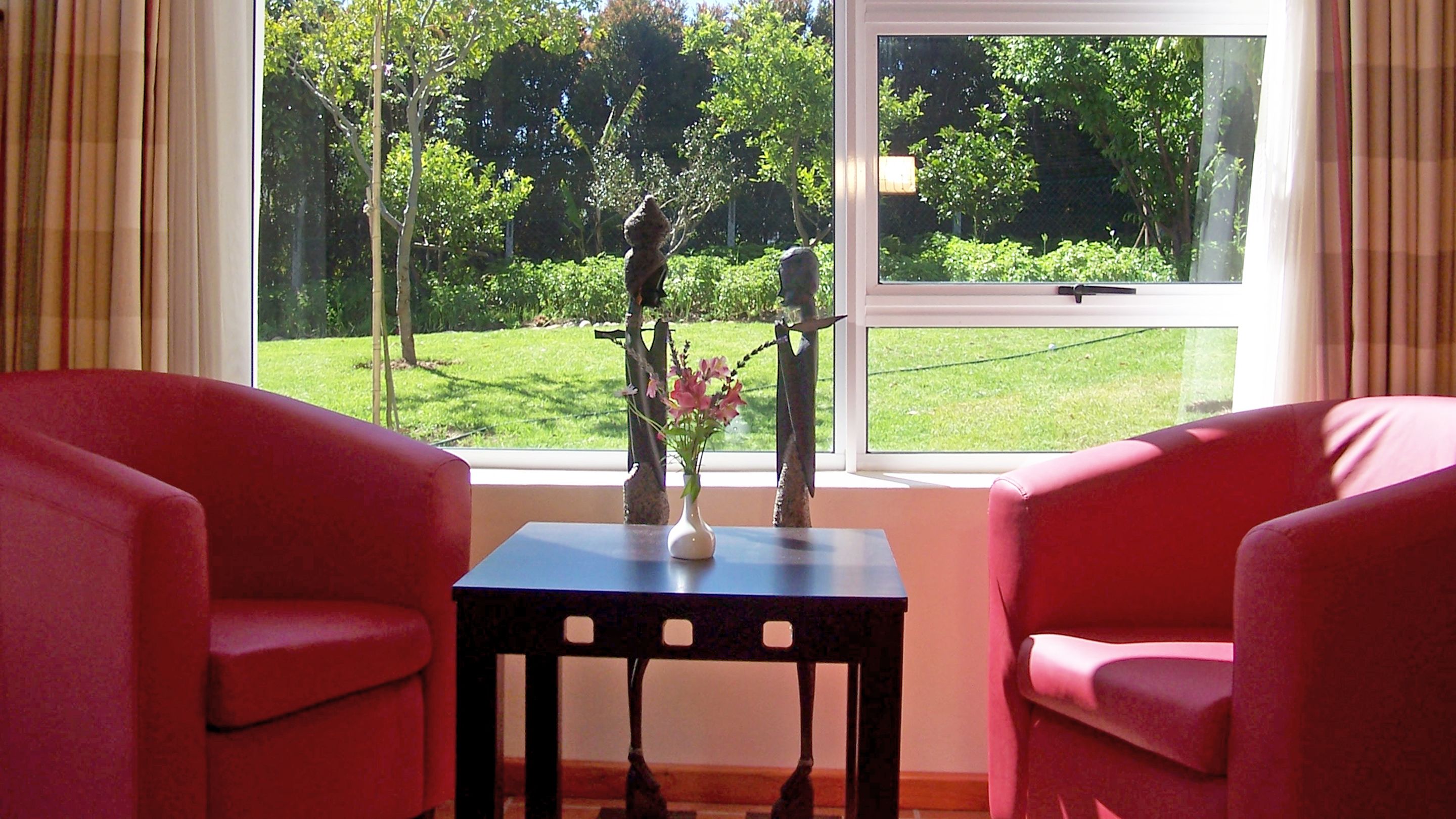 Una habitación con dos sillas rojas, una mesita con un florero y una ventana que da a un césped verde y árboles.