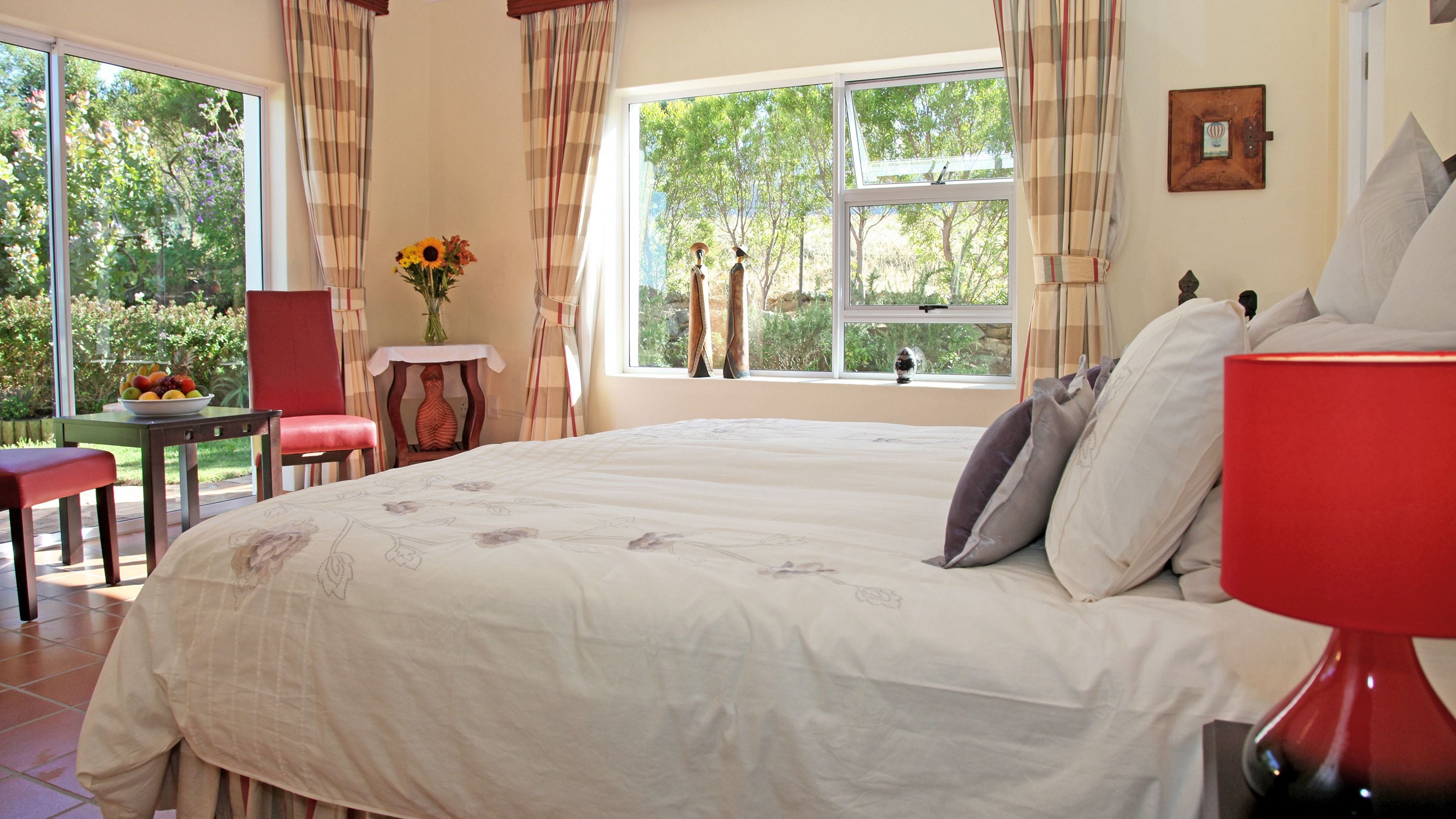 A bedroom with a large bed, a table with a vase of flowers, a chair, and a view of greenery outside.