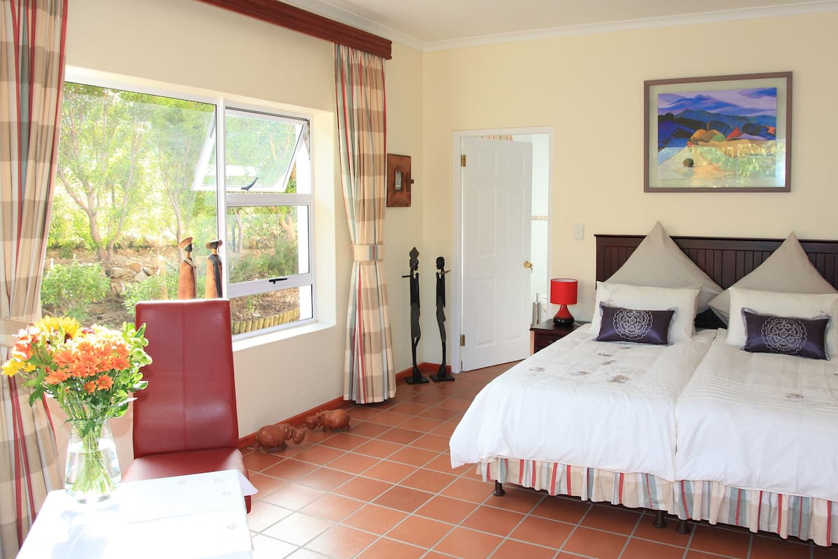 Une chambre avec un grand lit, deux tables de chevet, une fenêtre avec des rideaux, un tableau au mur et un vase rempli de fleurs sur une table.