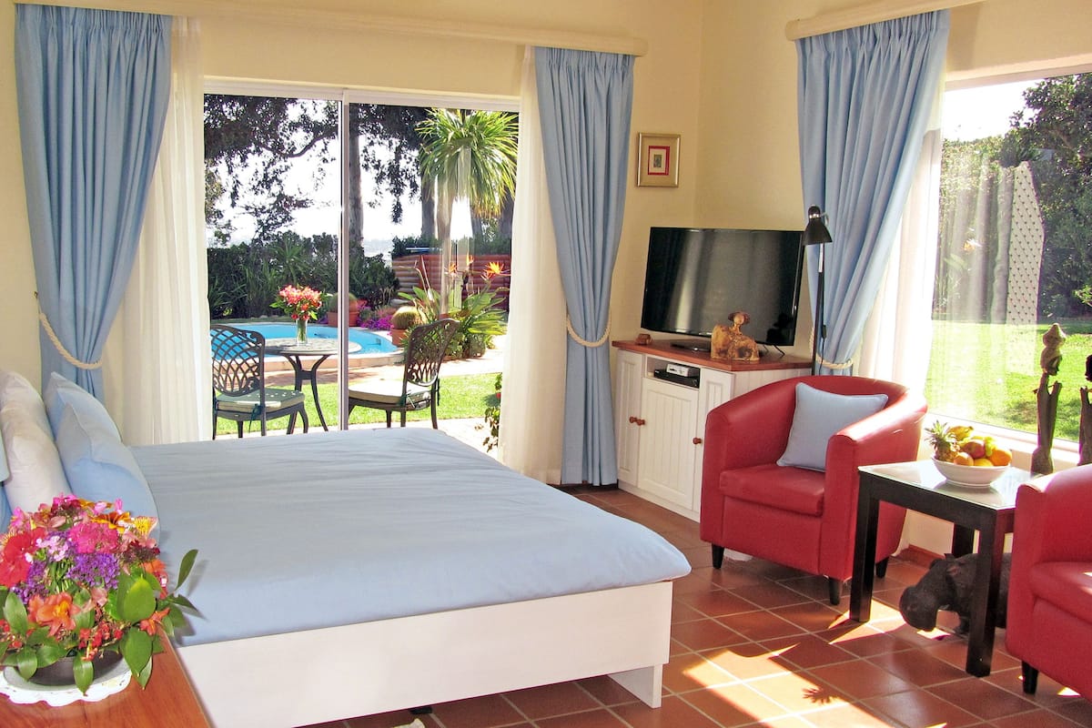 Une chambre avec un lit, une télévision, un bureau et une chaise. Il y a une fenêtre avec des rideaux, un vase avec des fleurs et une vue sur une piscine et un jardin.