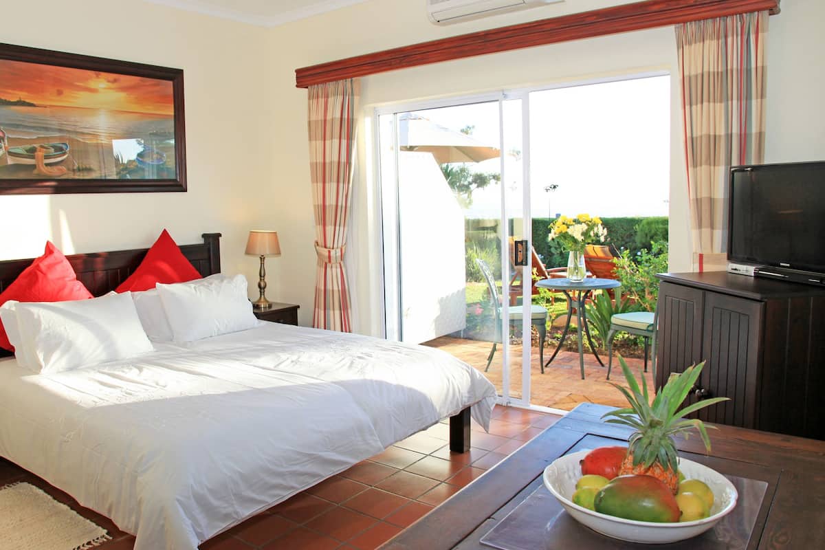 Une chambre d’hôtel avec un lit, une télévision, une table à manger avec des fruits et un tableau représentant un coucher de soleil sur l’océan.