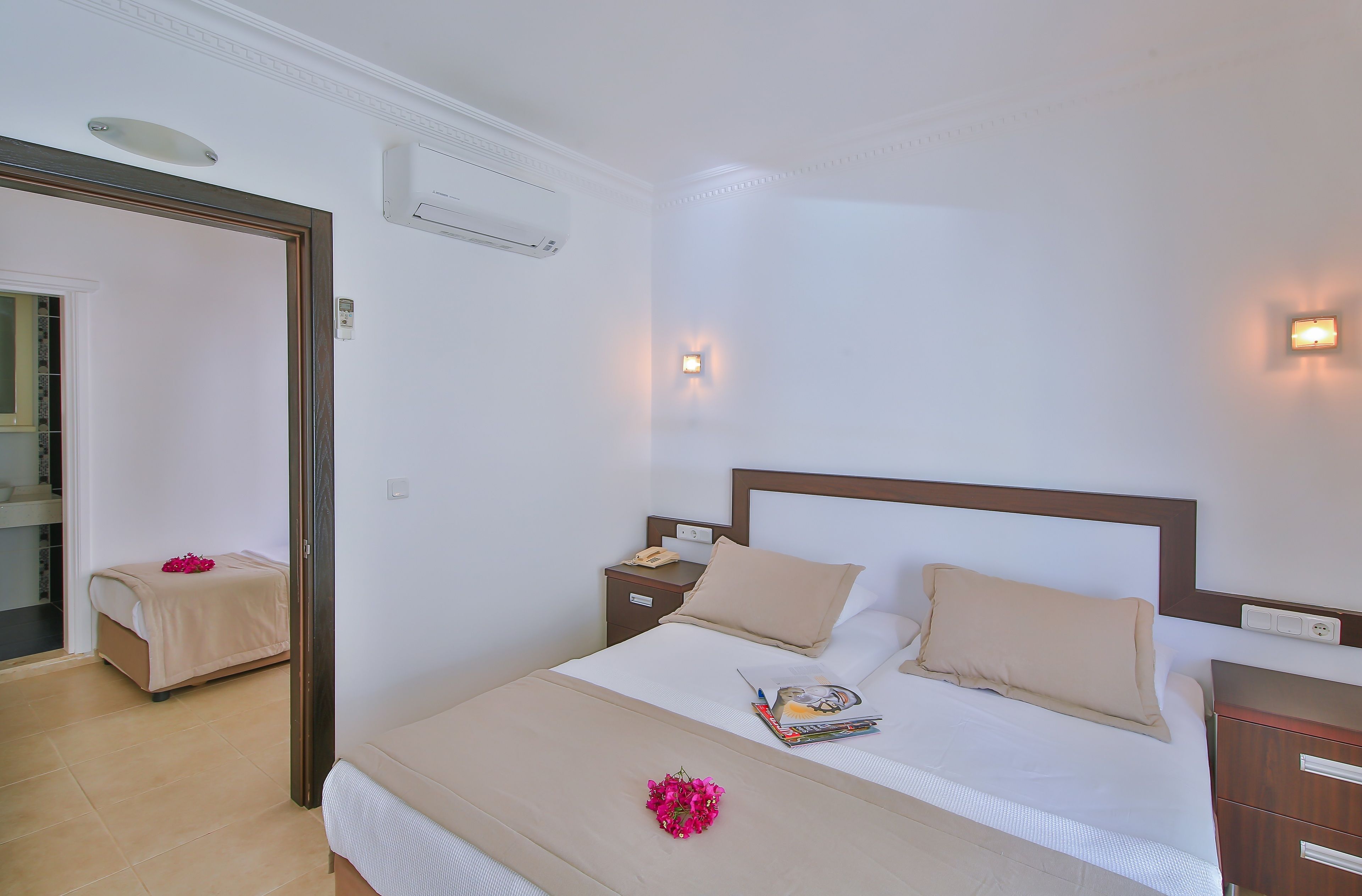 A neatly made bed with a beige bedspread, two pillows, and a magazine on the side. There is a nightstand with a phone and a small lamp. The room has white walls and a wooden headboard.