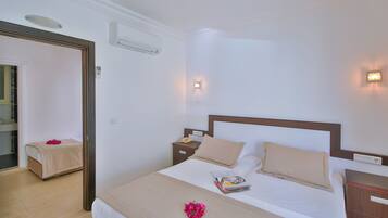 A neatly made bed with a beige bedspread, two pillows, and a magazine on the side. There is a nightstand with a phone and a small lamp. The room has white walls and a wooden headboard.