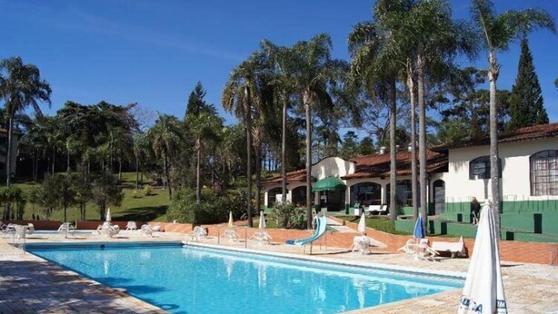 A swimming pool with lounge chairs and umbrellas, surrounded by palm trees and a building with a tiled roof. - Hotel Fazenda São Matheus (Serra Negra)