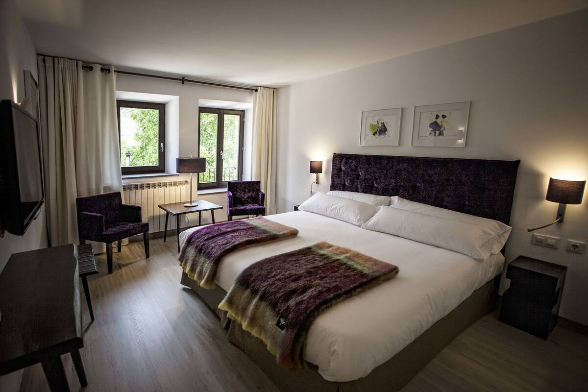 A hotel room with a large bed, two chairs, a desk, and a window with curtains.
