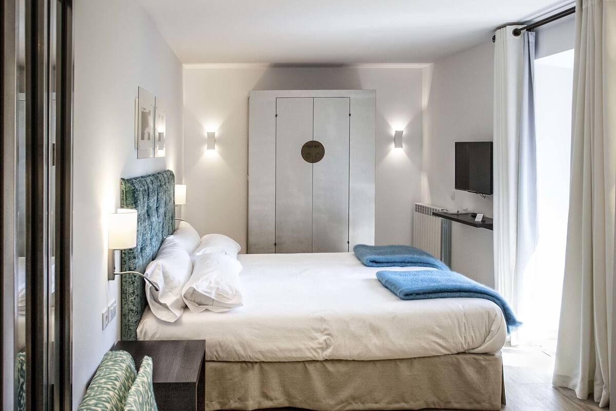 A hotel room with a bed, a seating area, a wardrobe, and a television.