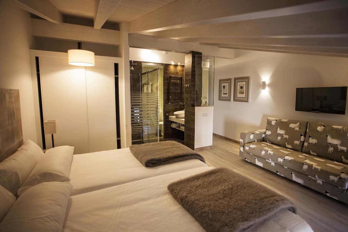 A modern hotel room with a bed, a sofa, a TV, and a bathroom visible through an open door.