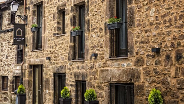 A stone building with multiple windows, some with plants. A sign indicates it is a hotel. - Hotel Pura Vida (Valganon)