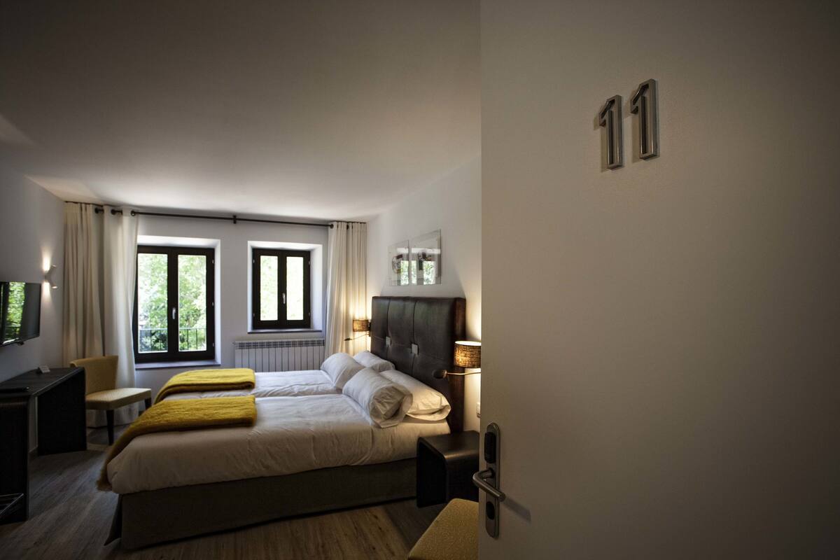 A hotel room with two beds, a desk, a chair, a window with curtains, and a wall-mounted TV.
