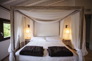 A four-poster bed with white linens and two dark throw pillows, flanked by nightstands with lamps, in a room with a wooden ceiling and a window with curtains. - Hotel Pura Vida (Valganon)