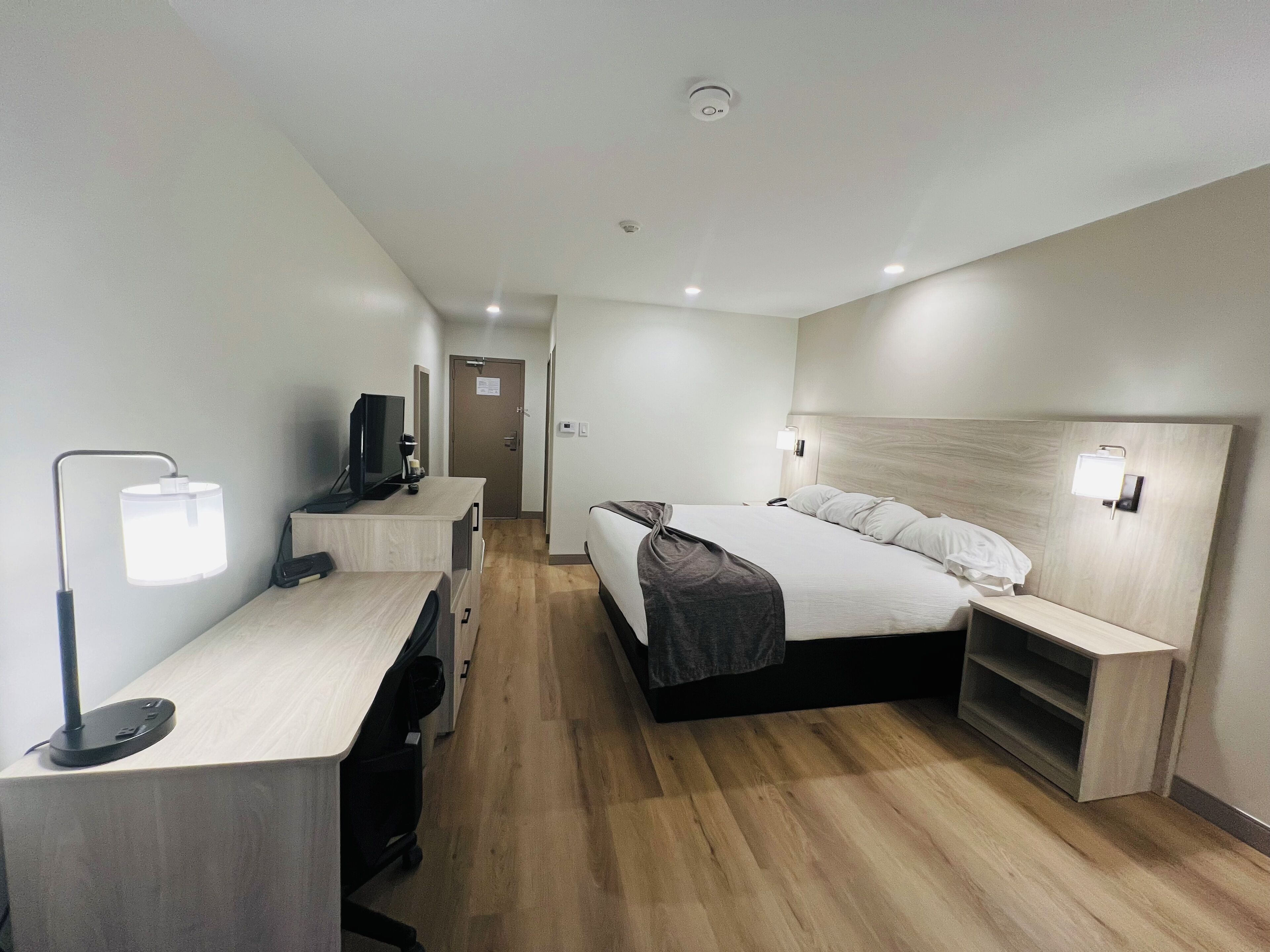 A modern hotel room with a wooden floor, a bed with white linens, a desk with a computer and lamp, and a television.