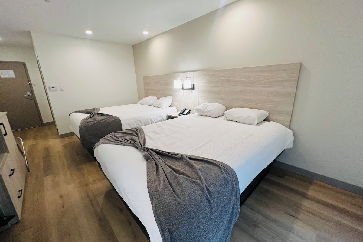 A hotel room with two beds, a wooden floor, a wall-mounted light, and a door with a sign.