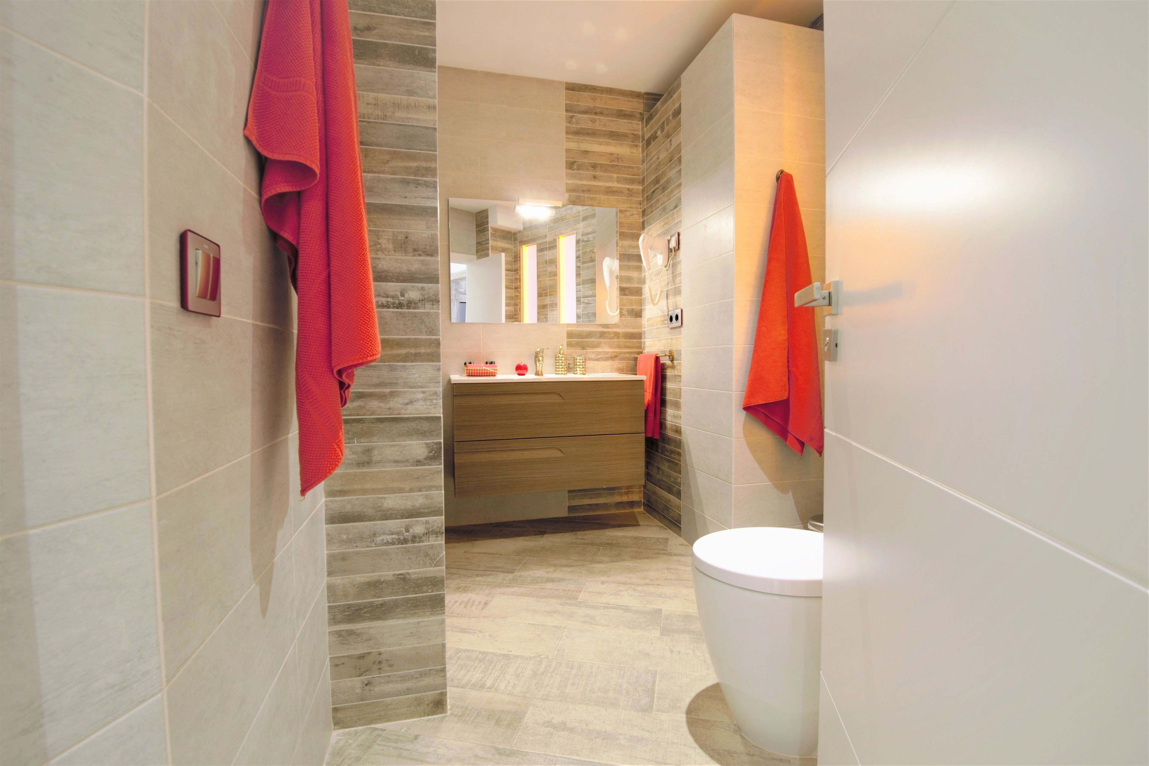 A modern bathroom with a wooden vanity, a white toilet, and red towels hanging on the walls.