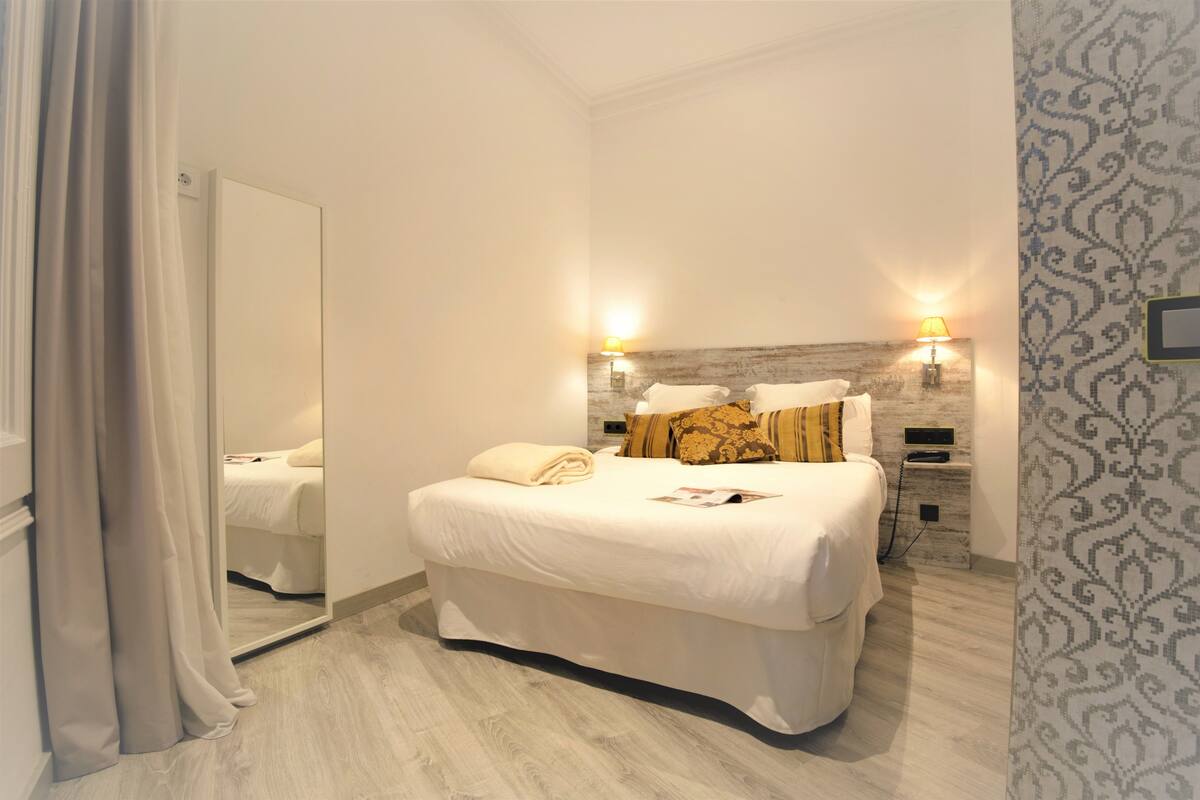 A neatly made bed with white linens, a mirror, two bedside lamps, and a patterned wall.