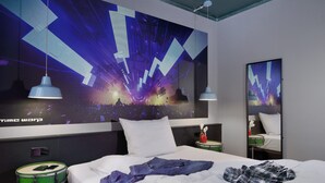 A modern hotel room with a large digital artwork on the wall, a neatly made bed, a green drum set, and a mirror reflecting part of the room. -  Staytion Urban City Hotel Mannheim (Mannheim)
