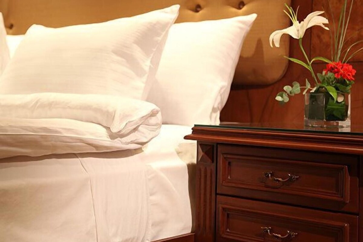 A bed with white linens, a wooden nightstand with drawers, and a vase with flowers.