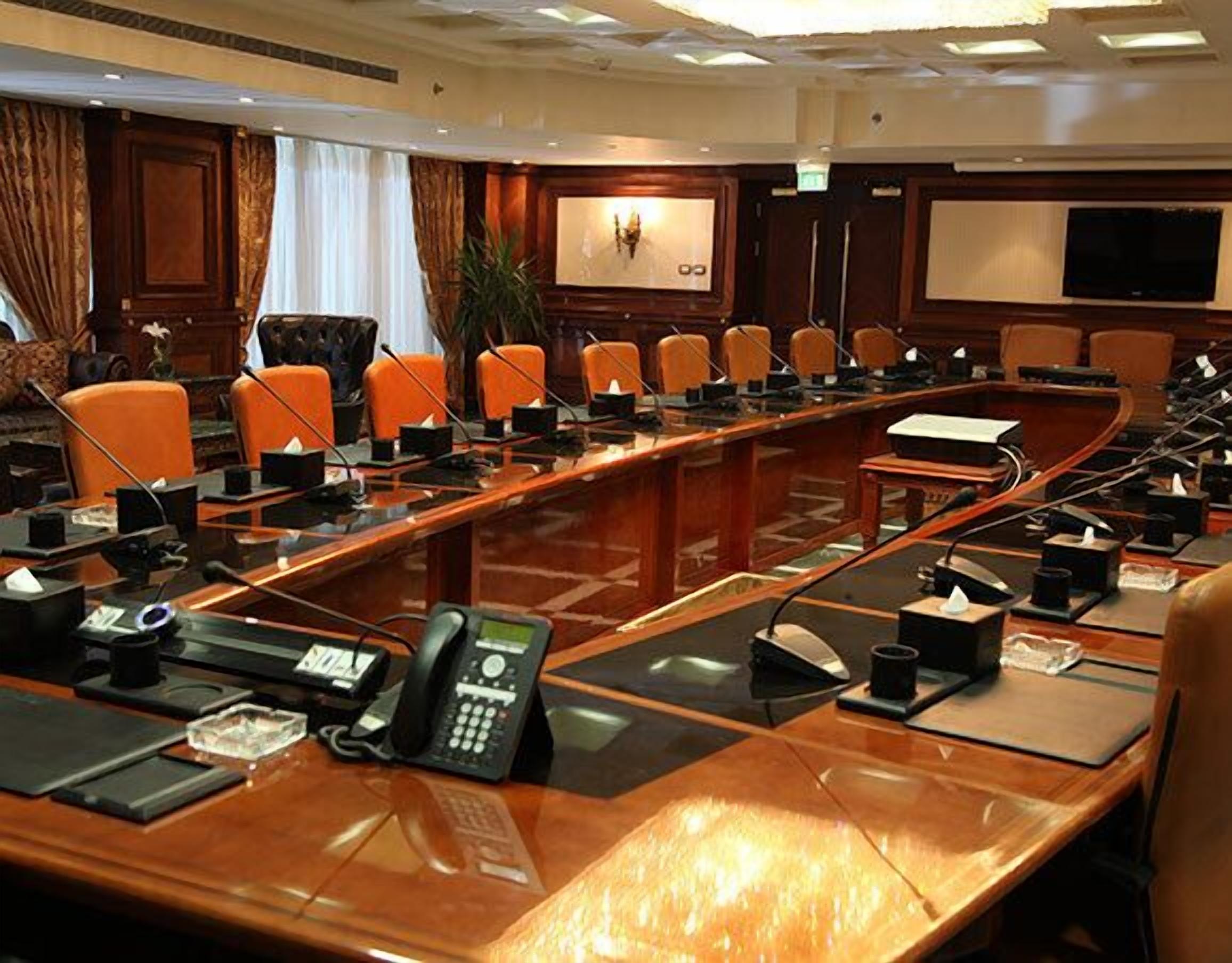 Un salón de conferencias con una larga mesa, varias sillas y una pantalla grande al frente.